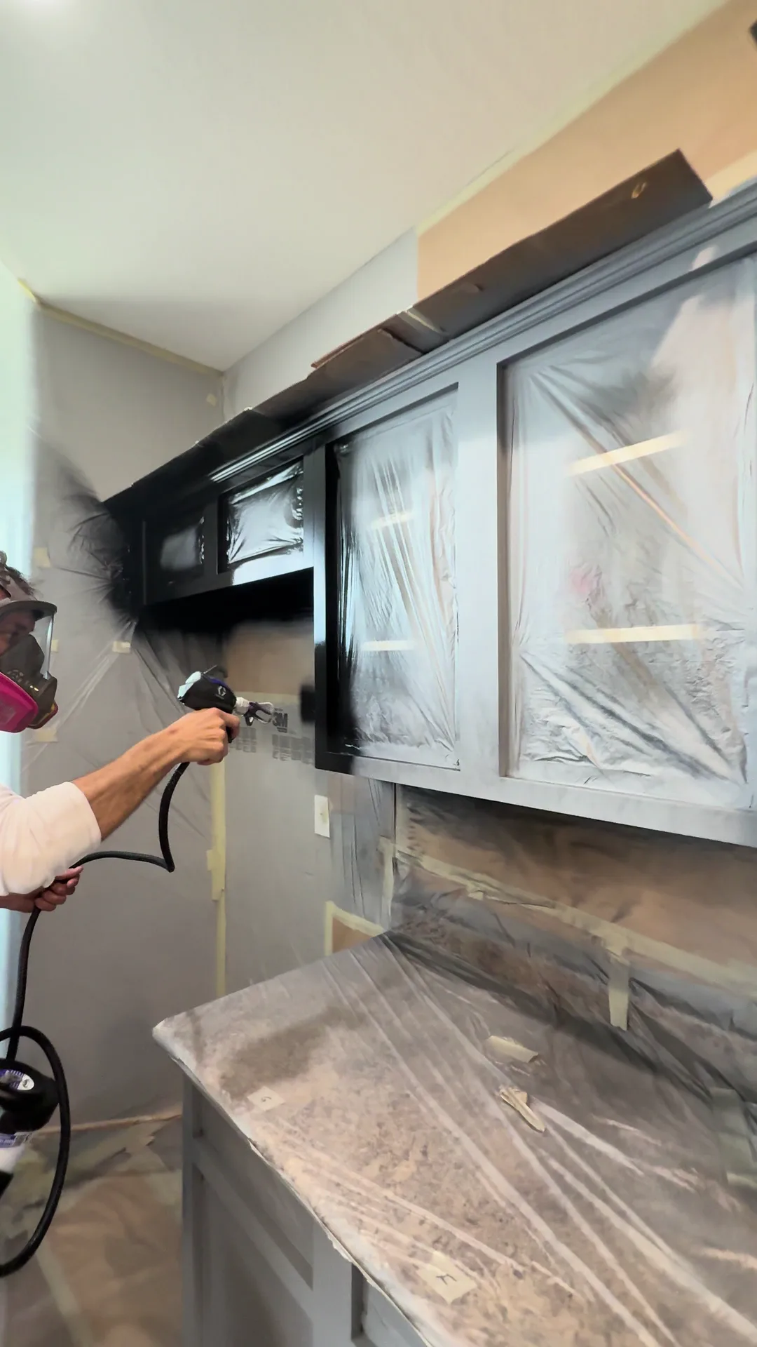 Ricardo Parra spraying the finish coat onto kitchen cabinets, in a respirator, with the counters and backsplash fully masked off.